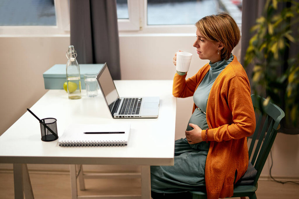 Красива біла вагітна бізнес-леді сидить у своєму кабінеті, торкається живота і п'є прісну воду
. - Фото, зображення