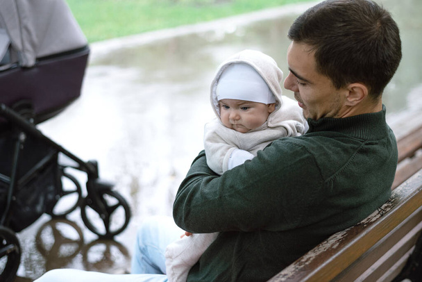 Kaukasische man vader in groene jas zitten op bank knuffelen zijn baby in Park - Foto, afbeelding