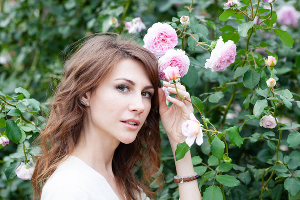 closeup portrait of beautiful young woman brunette with pink rose bush on background. Girl looks at camera. Her hand is close to hair - Foto, Imagem