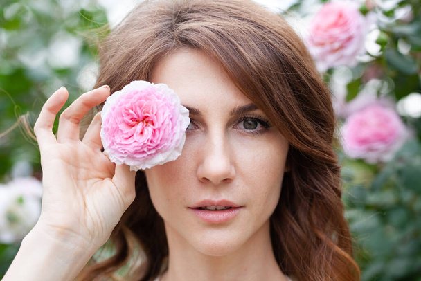 closeup portrait of beautiful young woman brunette with pink rose bush on background. Girl looks at camera and holds a flower in her hand, covering one eye with it - Foto, imagen
