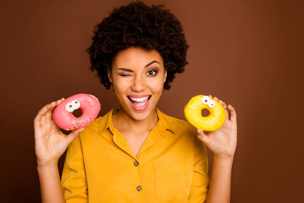 Photo of pretty dark skin wavy lady hold two colorful donuts caramel eyes human faces crazy carefree blinking flirty wear yellow shirt isolated brown color background - Photo, Image