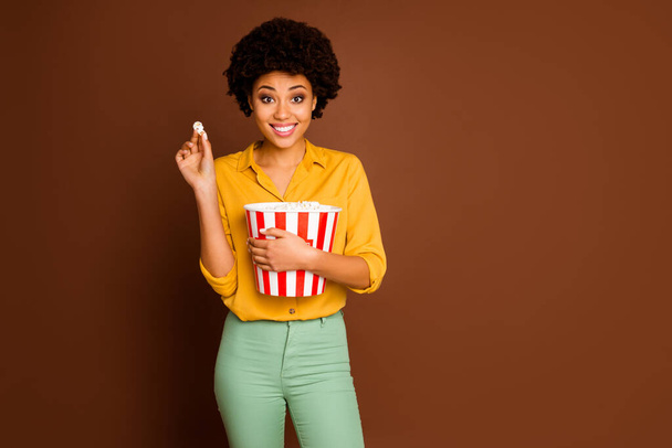 Photo of funny charming dark skin curly lady holding popcorn bucket eat corns watching favorite tv show wear yellow shirt green pants isolated brown color background - Φωτογραφία, εικόνα