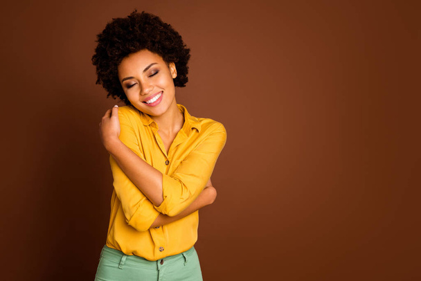 Photo of charming beautiful dark skin lady holding shoulders eyes closed enjoy love herself nice blouse clothes wear yellow shirt green trousers isolated brown color background - Фото, зображення