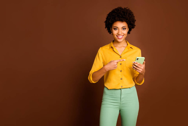 Portret pozytywnej wesołej afro amerykańskiej dziewczyny student używać smartfona wyszukiwania sprzedaży online wskazują punkt wskazujące urządzenie palca nosić stylowe żółte spodnie odizolowane brązowy kolor tła - Zdjęcie, obraz
