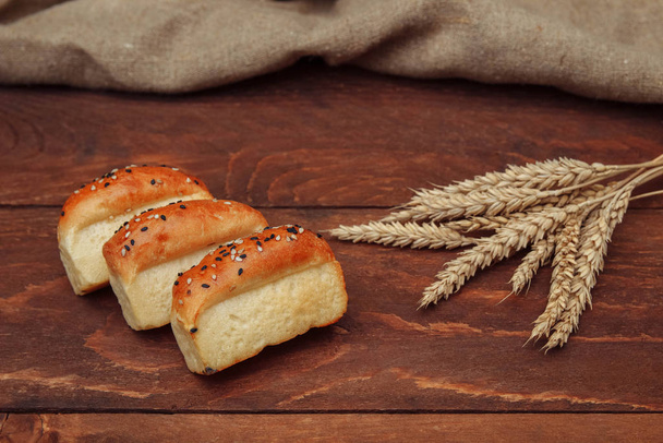 Three small baked bread rolls made from wheat flour, sprinkled with sesame seeds, lie on a brown wooden background with a bunch of wheat ears and a linen napkin - Photo, Image