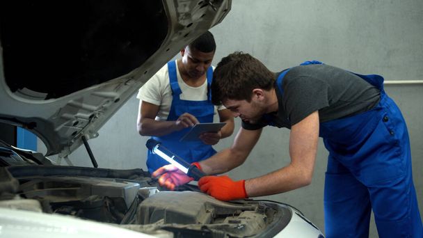 Μηχανικός με φακό εξετάζει τον κινητήρα του αυτοκινήτου και του collegue κρατά σημειώσεις - Φωτογραφία, εικόνα