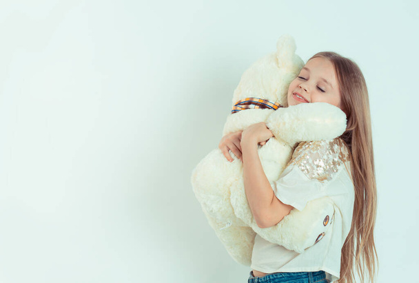 Cute happy kid girl with eyes closed hugging white teddy bear in studio. Closeup portrait of Caucasian kid model in white shirt isolated on white green gray copy space background. Horizontal image - Photo, image