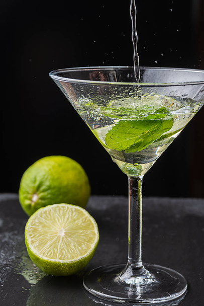 Close-up of cocktail glass with mint leaves and lime slice, pouring water, with splashes, on black background with a lime and a half with selective focus on vertical - Foto, Imagem
