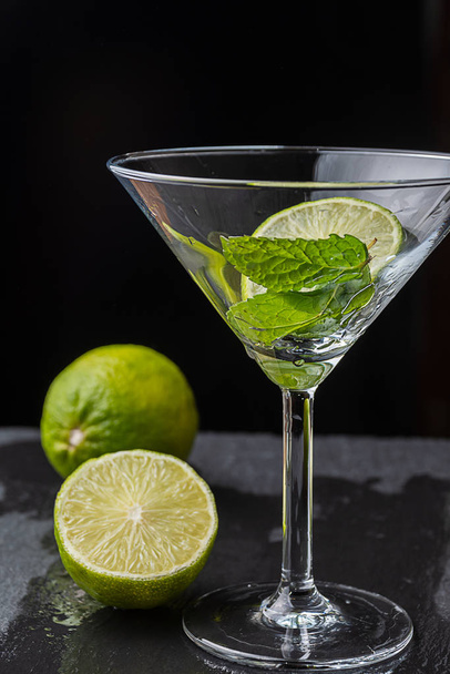 Close-up of cocktail glass with mint leaves and slice of lime on black background with a lime and a half with selective focus on vertical - Fotó, kép