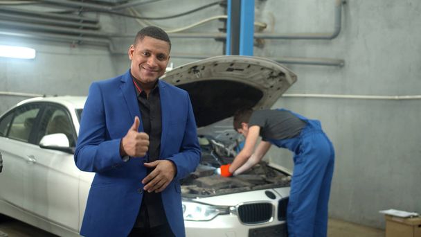 Медленное движение, человек улыбается и показывает хорошие признаки, механик ремонтирует автомобиль на заднем плане
 - Фото, изображение
