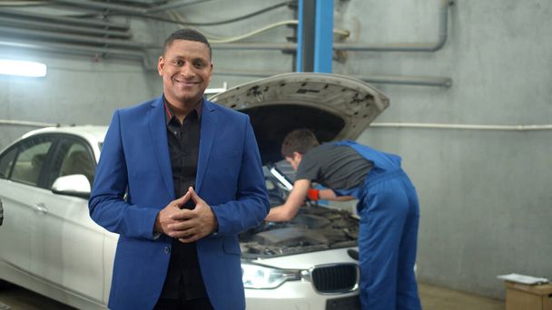 Egy öltönyös férfi mosolyog a kamerába, a szerelő megjavít egy autót a háttérben. - Fotó, kép