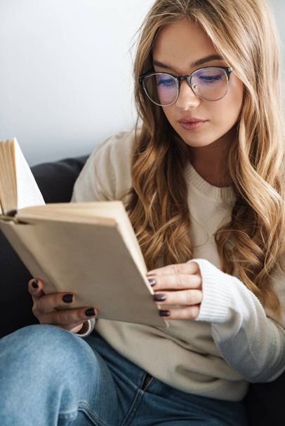 Photo of young serious woman reading book while sitting on couch - Foto, immagini