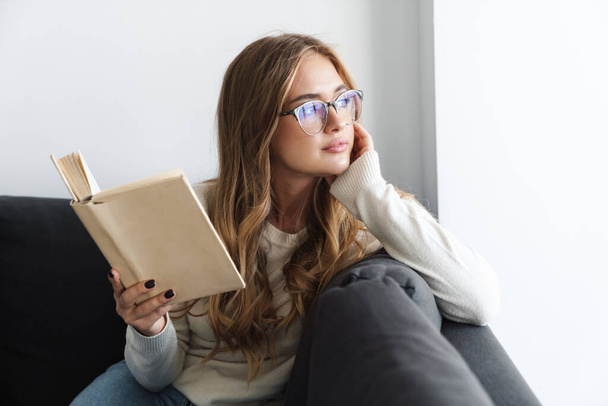 Photo of young serious woman reading book while sitting on couch - Fotó, kép