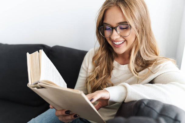 Photo of young smiling woman reading book while sitting on couch - Fotó, kép