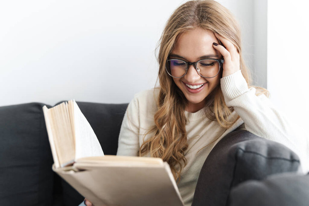Photo of young smiling woman reading book while sitting on couch - Foto, Imagem