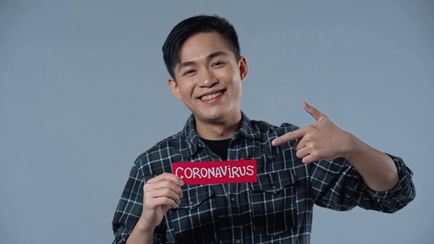 glücklicher asiatischer Mann zeigt mit dem Finger auf Coronasvirus Schriftzug isoliert auf grau  - Filmmaterial, Video