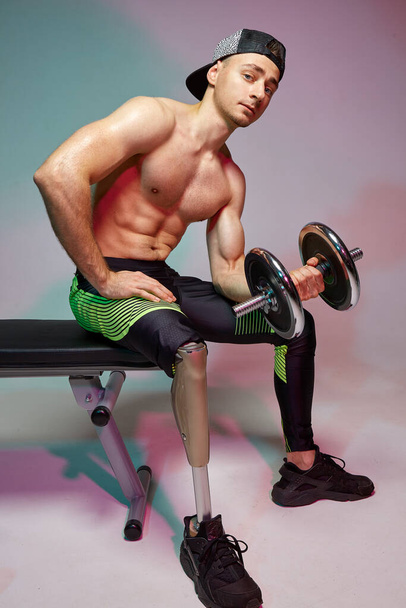 A young muscular man with a disability with a prosthetic leg is training in a gym with dumbbells. Motivation poster. - Fotoğraf, Görsel
