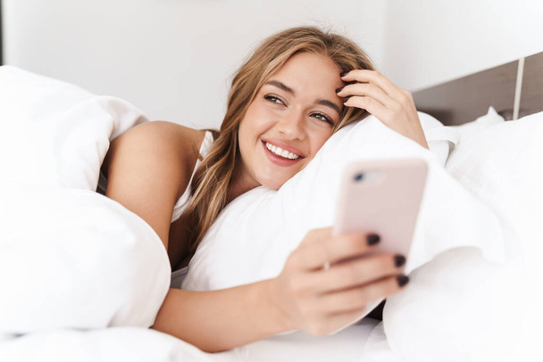 Photo of smiling caucasian woman using cellphone while lying in bed - Foto, immagini