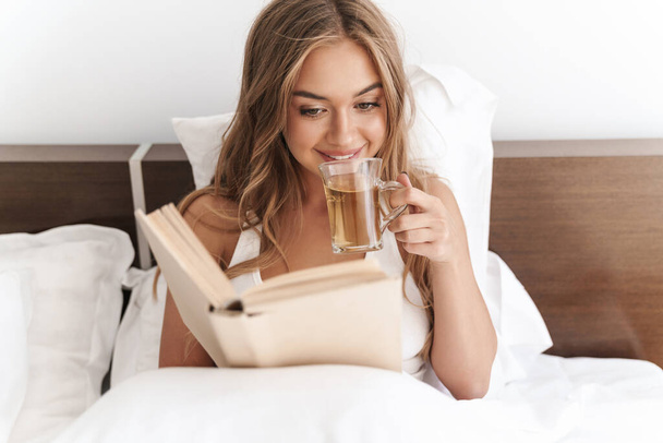 Photo of young joyful woman reading book and drinking tea - Foto, Imagen