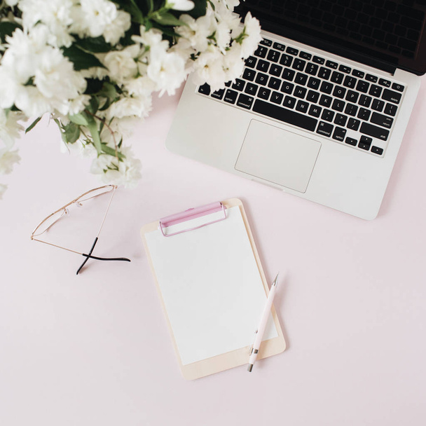 Office desk workspace with laptop, flowers bouquet and blank paper copy space mockup clipboard on pink table. Flat lat, top view freelancer co-working background. - Foto, Bild