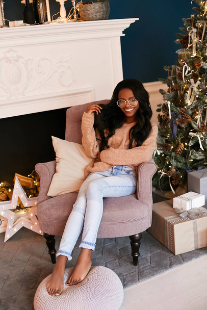Glamorous black girl in a luxurious Christmas interior sits in a stylish armchair in the room. Decorative fireplace and Christmas tree in the background. - Foto, Bild