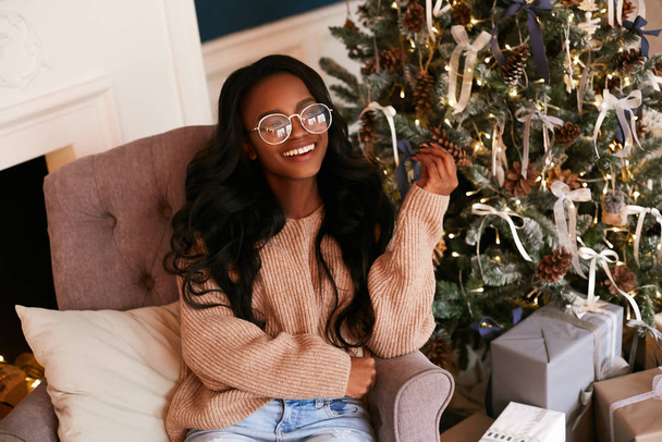 Happy fashionable black girl in a luxurious Christmas interior. African American woman sitting in a stylish armchair against the background of the Christmas tree. - Fotografie, Obrázek
