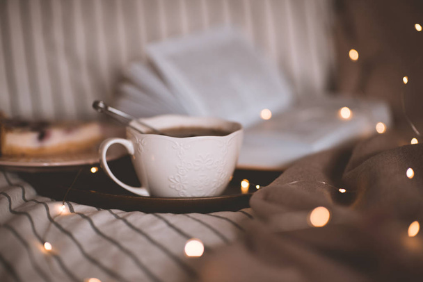 Taza de té negro fresco con libro abierto en primer plano de la cama sobre luces brillantes en el fondo. Buenos días. Hora del desayuno
.  - Foto, Imagen