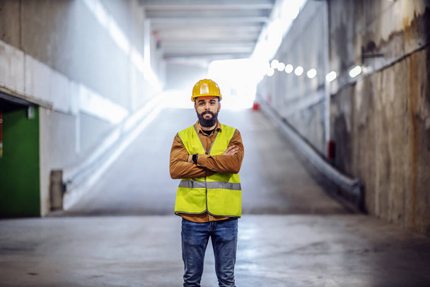 Молодой привлекательный успешный бородатый супервайзер, стоящий у входа на подземную автостоянку в процессе строительства со сложенными руками
. - Фото, изображение