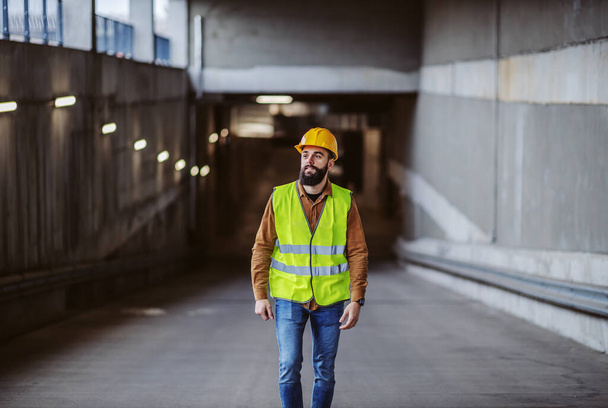 Полная длина трудолюбивого, привлекательного бородатого работника в жилете, с защитным шлемом на голове, выходящим из здания в строительной зоне
. - Фото, изображение