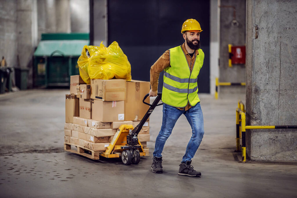Full length of smiling hardworking bearded worker in vest, with safety helmet on head pulling a pallet truck with boxes, sack and building material. Building in construction process interior. - Foto, Bild