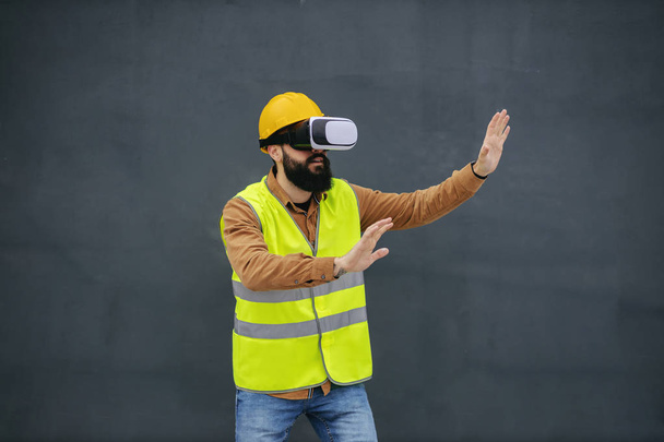 Молодой бородатый рабочий в жилете, с защитным шлемом на голове, в очках и наслаждается виртуальной презентацией
. - Фото, изображение