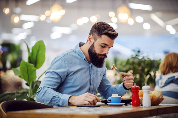 Привлекательный голодный бородатый хипстер сидит в ресторане и ест свой завтрак
. - Фото, изображение