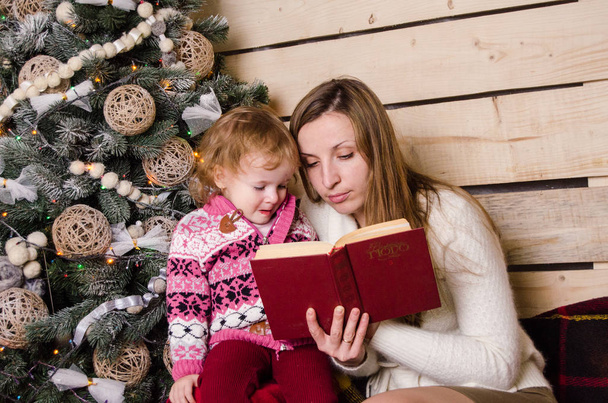 gyönyörű fiatal anya és lánya olvasás könyv közelében díszített karácsonyfa - Fotó, kép