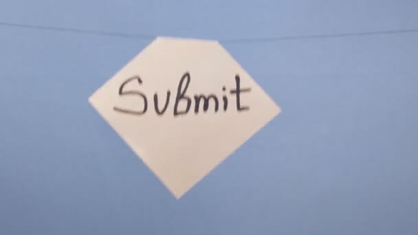 Um homem pendura uma folha branca de papel com uma inscrição preta "submeter" em um fundo azul
 - Filmagem, Vídeo