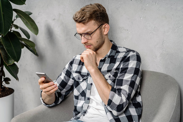 Nuori aikuinen mies käyttää modernia älypuhelinta, istuu nojatuolissa
 - Valokuva, kuva