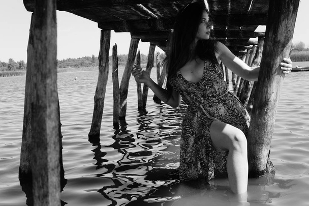 Atractiva mujer rubia sentada en el muelle junto al lago con fondo natural. Novia romántica con vestido blanco y ramo de flores
. - Foto, Imagen