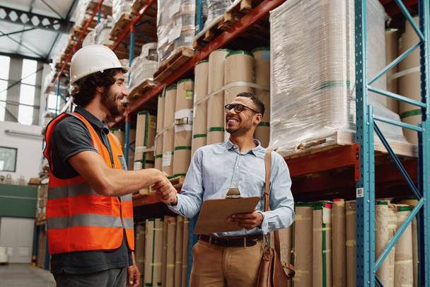 Cheerful young business man in formal clothing wearing side bag and holding clipboard making handshake gesture with warehouse manager - two business partners making a deal  - Foto, imagen