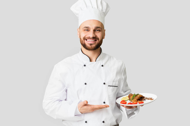 Uśmiechnięty kucharz obsługujący danie z kurczaka stojący na szarym tle - Zdjęcie, obraz