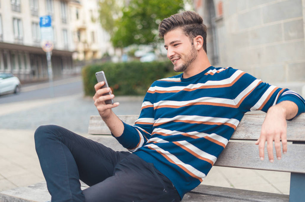Side view of a happy guy texting on a smart phone while sitting on the bench - 写真・画像