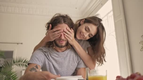 Happy pretty woman covering eyes her boyfriend while man reading book and sitting by the table on kitchen at home - Imágenes, Vídeo