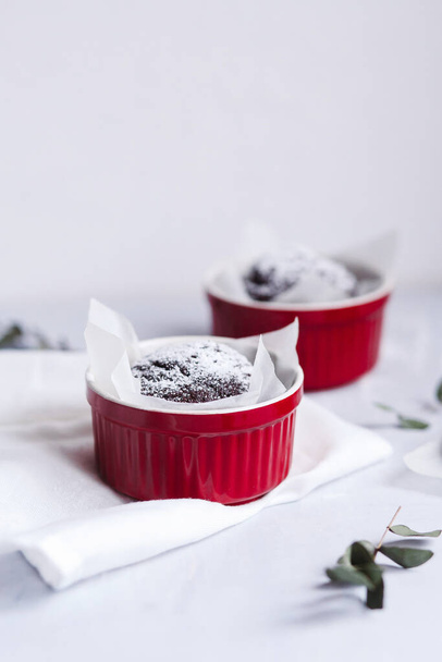 Muffins au chocolat dans des tasses rouges. Petit ramequin en céramique émaillée avec des gâteaux bruns sur fond gris et blanc
. - Photo, image