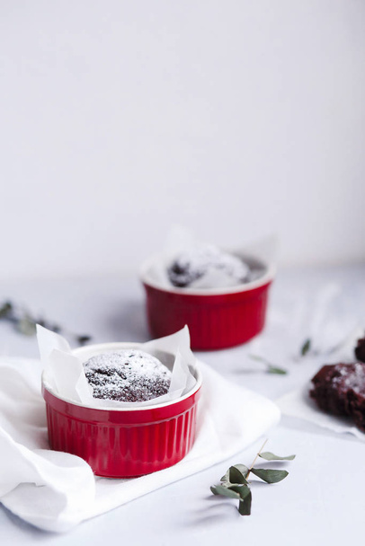 Chocolate muffins in red cups. Small glazed ceramic ramekin with brown cakes on a gray and white background. - 写真・画像
