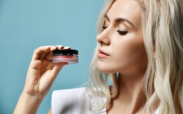 Young beautiful sexy blond woman cosmetologist in white medical uniform is holding looking at skin cream small jar - Foto, immagini