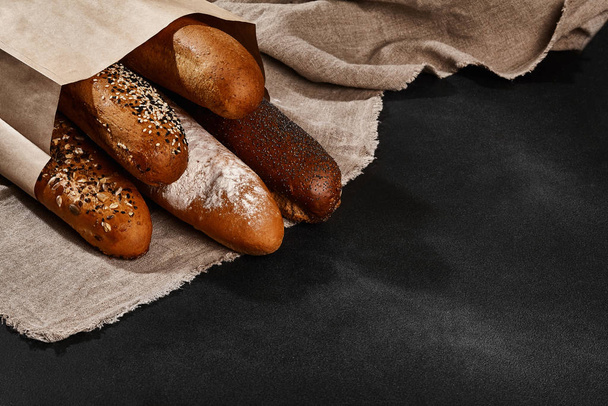 Verscheidenheid van vers gebakken brood in een papieren zak, liggend op een jute tegen zwarte achtergrond, kopieer ruimte. Close-up - Foto, afbeelding