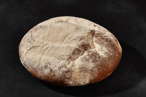 Świeży, pyszny pieczony okrągły chleb posypany mąką. Czarny tło z kopia przestrzeń. Kuchnia wiejska lub piekarnia. Zbliżenie - Zdjęcie, obraz