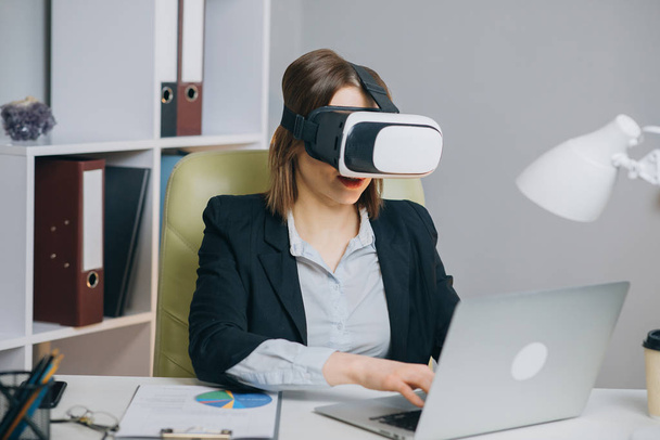 Woman wearing virtual reality glasses in office. Young girl using VR goggles in office. Pretty woman working in augmented reality at workplace. - 写真・画像