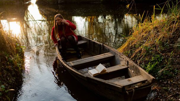 göndör lány ül a szélén egy elhagyatott fa csónak naplementekor. - Fotó, kép