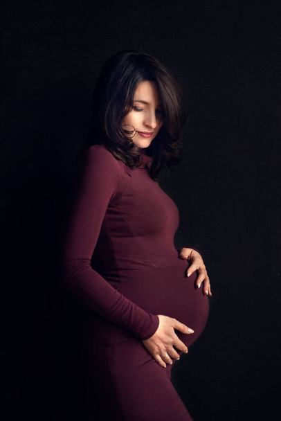 Pregnant happy Woman touching her belly. Pregnant middle aged mother portrait, caressing her belly and smiling close-up. Healthy Pregnancy concept, brunette expectant female on black background - Φωτογραφία, εικόνα