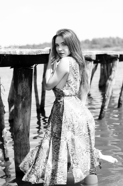 Dark-haired woman posing at the wooden pier at the lake. Woman in summer outfit with her back turned to the wooden footbridge of Lake Neusiedl. Sun reflections at the boathouse in the background - Photo, Image