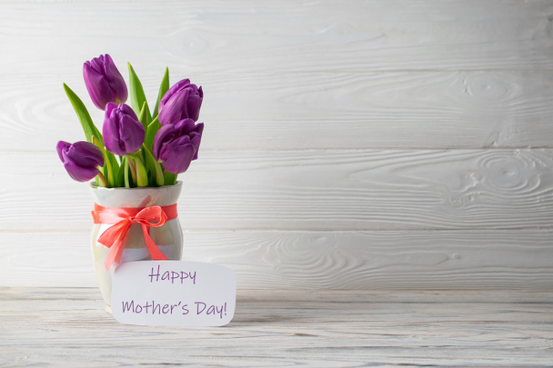 Mother's Day holiday card with a bouquet of fresh purple tulips in a vase with a pink bow on a white wooden background. Flat lay with copy space for you text - Photo, Image